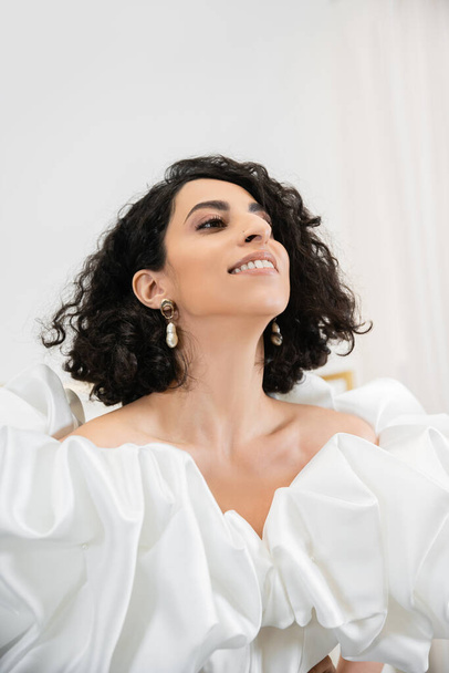 portrait of happy middle eastern bride with brunette and wavy hair posing in trendy wedding dress with puff sleeves and ruffles looking up in bridal salon, white gown - Fotografie, Obrázek