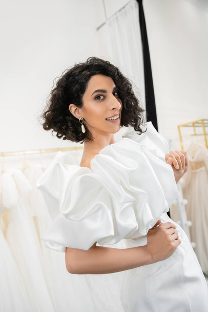 happy middle eastern bride with brunette and wavy hair posing with hands on hips in trendy wedding dress with puff sleeves and ruffles in bridal boutique next to tulle fabrics  - Φωτογραφία, εικόνα