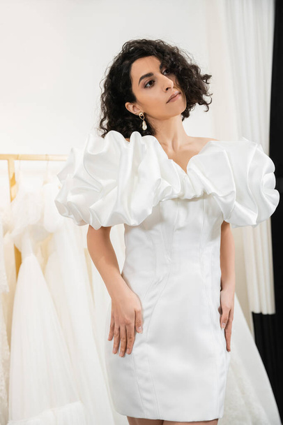 elegant middle eastern bride with brunette and wavy hair posing in trendy wedding dress with puff sleeves and ruffles in bridal boutique next to tulle fabrics, elegant woman  - Zdjęcie, obraz