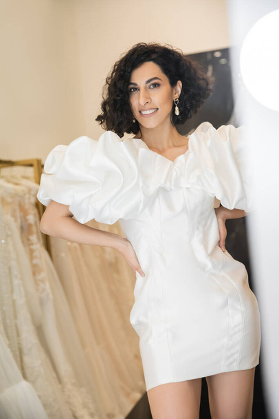 happy middle eastern woman with brunette wavy hair trying on wedding dress with puff sleeves and ruffles near mirror in bridal boutique next to tulle fabrics, reflection, shopping, hands on hips - Photo, Image