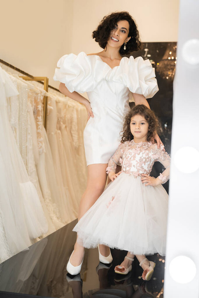joyful middle eastern bride with wavy hair posing in trendy wedding dress with puff sleeves and ruffles near cute little daughter in floral attire and white bridal gown in boutique   - Photo, Image
