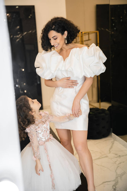 happy and cute middle eastern little girl in floral attire looking at bride with wavy hair standing in trendy wedding dress with puff sleeves and ruffles in bridal boutique, mother and daughter  - Photo, Image