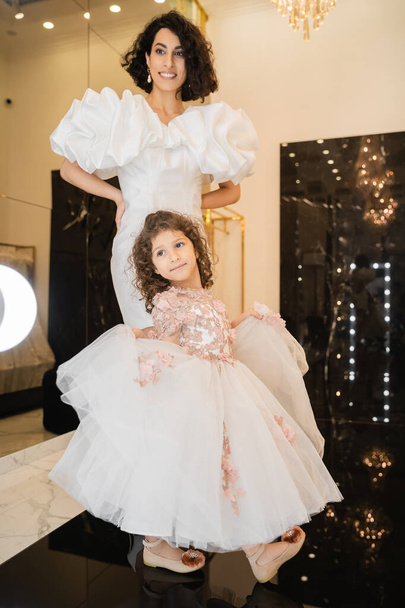 charming middle eastern bride with brunette hair standing in white wedding gown with puff sleeves and ruffles and looking at mirror near daughter holding tulle skirt in bridal store  - Foto, Imagem