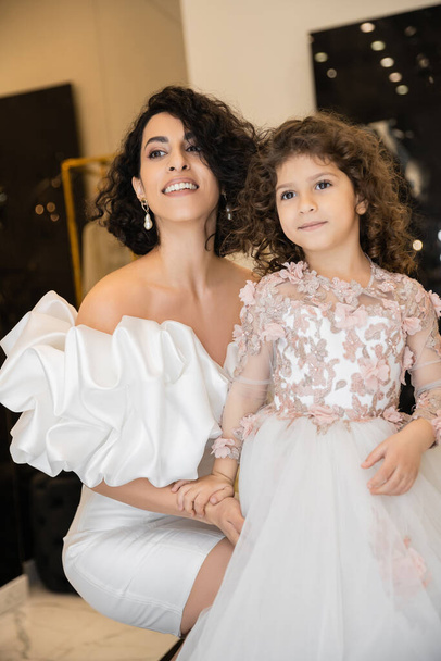 enchanting middle eastern bride with brunette hair in white wedding dress with puff sleeves and ruffles hugging daughter and looking away in bridal store, special occasion  - Photo, Image