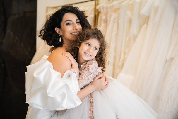 charming middle eastern bride with brunette hair in white wedding dress with puff sleeves and ruffles embracing positive daughter in bridal store, white tulle fabrics on blurred background  - Photo, Image