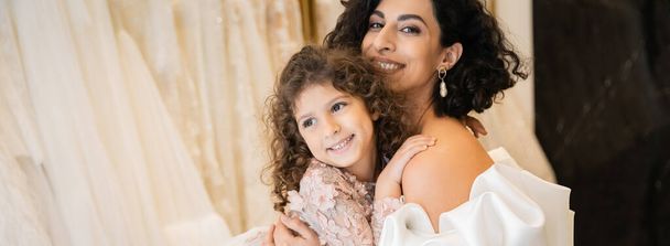 gorgeous middle eastern bride with brunette hair in white wedding dress with puff sleeves and ruffles embracing positive daughter in bridal store, white tulle fabrics on blurred background, banner  - Photo, Image