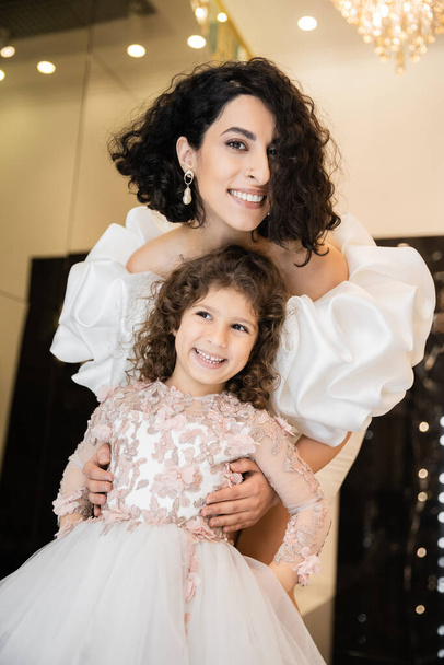 gorgeous middle eastern bride with brunette hair standing in white wedding dress with puff sleeves and ruffles behind cute daughter and smiling together in bridal store, looking at camera  - Photo, Image