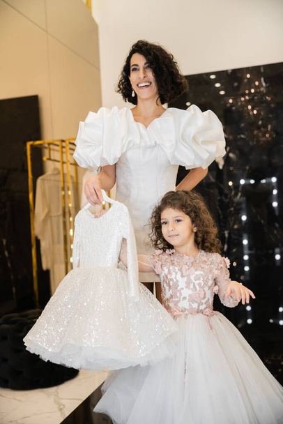 charming middle eastern bride with brunette hair standing in white wedding gown with puff sleeves and ruffles and holding girly dress with tulle skirt near daughter in bridal store  - Photo, Image