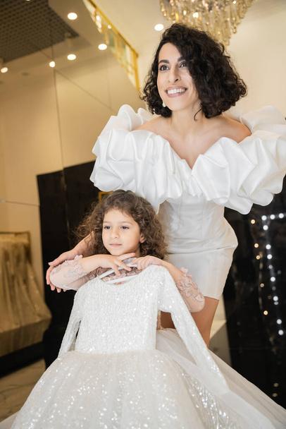 joyous middle eastern bride with brunette hair standing in white wedding gown with puff sleeves and ruffles near cute daughter holding girly dress with tulle skirt in bridal boutique   - Photo, Image