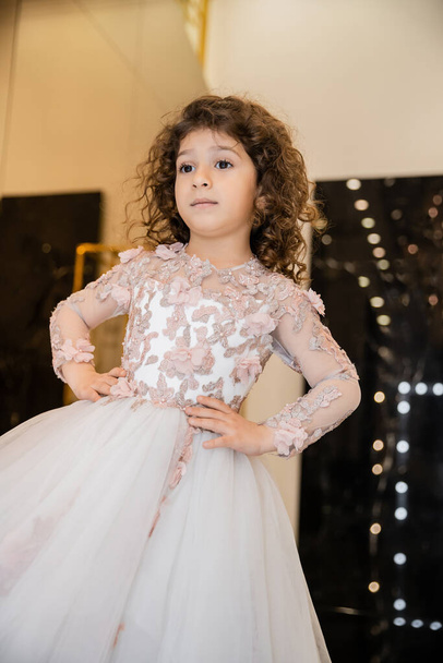 curly middle eastern and little girl in floral dress with tulle skirt standing with hands on hips and looking away in bridal boutique, preparation for wedding, blurred background, golden accents - Photo, Image