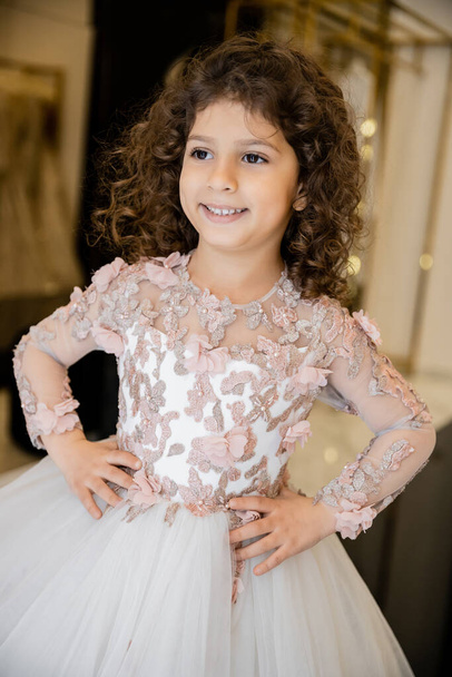 joyous middle eastern and little girl in floral dress with tulle skirt standing with hands on hips and looking away in bridal boutique, preparation for wedding, blurred background  - Photo, Image