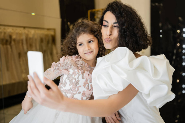 mittelöstliche Braut mit brünetten Haaren in weißem Brautkleid mit Puffärmeln und Rüschen macht Selfie auf Smartphone, während schmollende Lippen neben glücklicher Tochter im Brautladen  - Foto, Bild