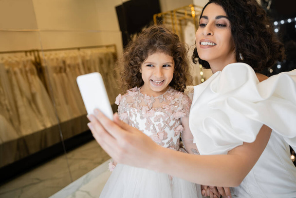 charming middle eastern bride with brunette hair in white wedding dress with puff sleeves and ruffles taking selfie on smartphone with happy daughter in bridal store with blurred background  - Photo, Image