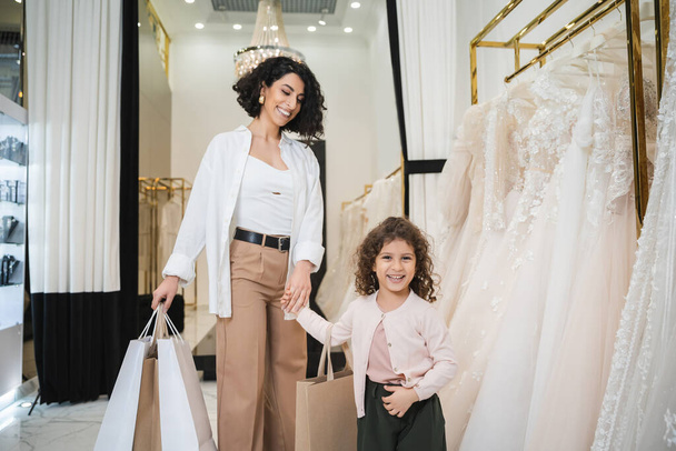 happy middle eastern bride with brunette hair in beige pants with white shirt holding shopping bags while standing with little girl near wedding dresses in bridal salon, mother and daughter  - Photo, Image