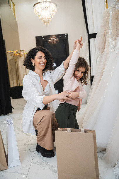 χαρούμενη γυναίκα της Μέσης Ανατολής με καστανά μαλλιά σε λευκό πουκάμισο κάθεται κοντά σε τσάντες για ψώνια και κρατώντας τα χέρια του μικρού κοριτσιού που χορεύει κοντά σε νυφικά φορέματα στο σαλόνι, μητέρα και κόρη, νύφη  - Φωτογραφία, εικόνα