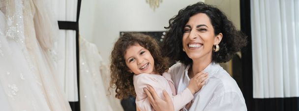 curly little girl hugging cheerful middle eastern bride with brunette hair in white shirt sitting near white wedding dresses in bridal salon, mother and daughter, bonding, special moment, banner  - Photo, Image