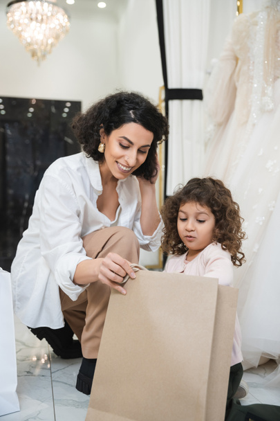 cheerful middle eastern woman with brunette hair in white shirt and surprised little girl looking inside of shopping bag near white wedding dresses in bridal salon, mother and daughter  - Foto, Bild