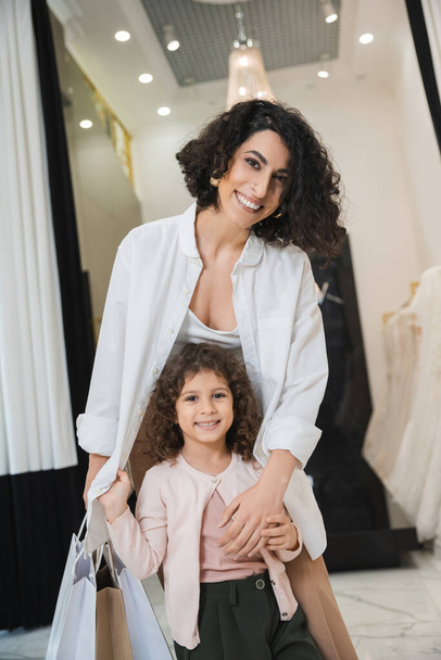 fröhliche Frau aus dem Mittleren Osten mit brünetten Haaren, die Einkaufstaschen hält und niedliches kleines Mädchen umarmt, während sie neben Brautkleidern im Brautsalon steht, Mutter und Tochter, Brauteinkäufe  - Foto, Bild