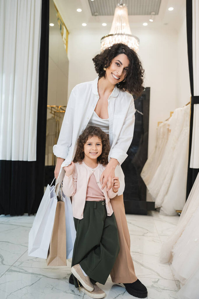 joyful middle eastern woman with brunette hair holding shopping bags and hugging cute little girl while standing near white wedding dresses in bridal salon, mother and daughter, bridal shopping  - Photo, Image