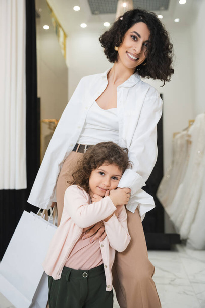 cute little girl hugging hand of happy middle eastern woman with brunette hair holding shopping bags while standing near wedding dresses in bridal salon, mother and daughter, bridal shopping  - 写真・画像