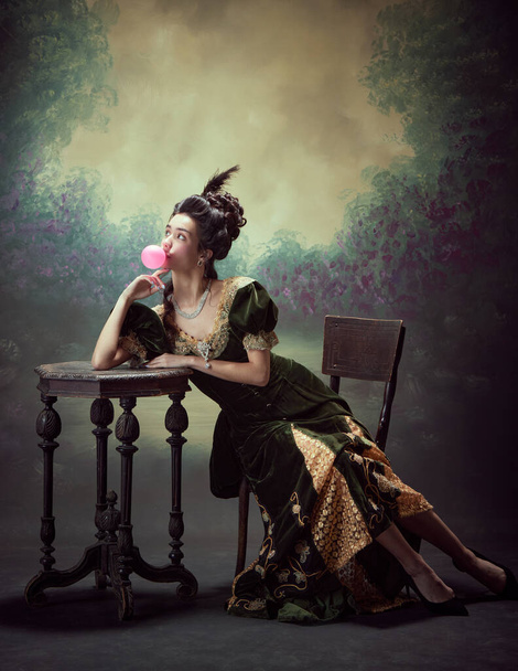 Portrait de jolie jeune fille, princesse élégante, personnage royal médiéval en robe rétro, mangeant du bubblegum sur fond vintage sombre. Concept d'histoire, art de la renaissance, comparaison des époques - Photo, image