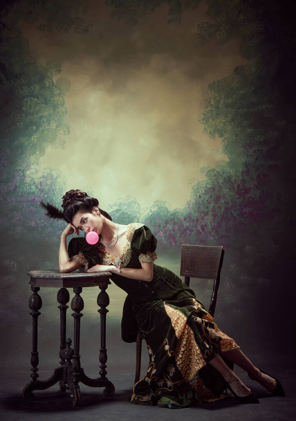 Portrait of sad young girl, elegant princess, medieval royal person in retro dress, eating bubblegum against dark vintage background. Concept of history, renaissance art, comparison of eras - Photo, image