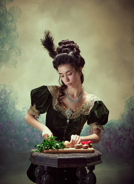 Portrait of tender young girl, elegant princess in stylish medieval dress cooking, cutting vegetables against dark vintage background. Concept of history, renaissance art, comparison of eras - Foto, imagen