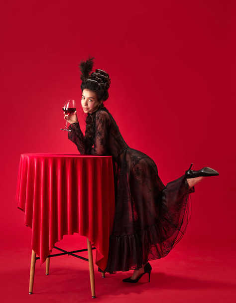 Portrait of beautiful, attractive young girl in elegant, stylish, black dress leaning on table, holding glass with red wine against red background. History, renaissance art, comparison of eras concept - 写真・画像