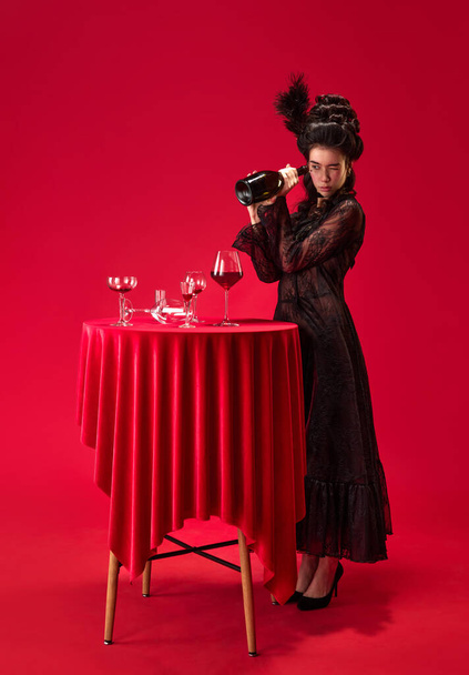 Time to drink. Portrait of beautiful, emotive young girl in elegant, stylish, black dress looking into wine bottle against red background. Concept of history, renaissance art, comparison of eras - Fotó, kép