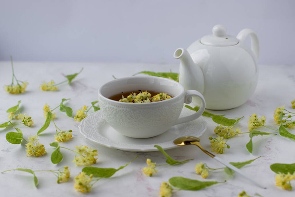 Τσάι με linden σε λευκό κύπελλο σε λευκό μαρμάρινο τραπέζι. Λευκή τσαγιέρα με αρωματικό τσάι - Φωτογραφία, εικόνα