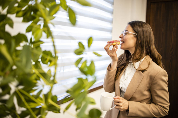 Красивая женщина наслаждается моментом и ест вкусные пончики на работе. - Фото, изображение