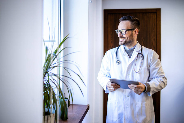 Опытный врач в белой форме и стетоскопе смотрит в окно больницы. - Фото, изображение