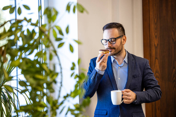 Homme d'affaires en costume élégant ayant une pause café et manger de délicieux beignets à l'intérieur du bureau. - Photo, image