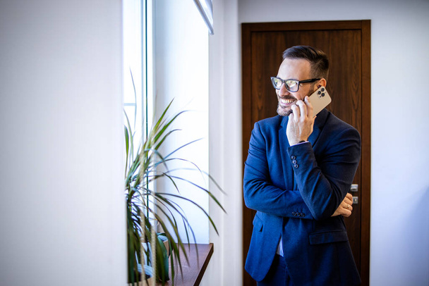 Podnikatel stojí u okna kanceláře a mluví do telefonu. - Fotografie, Obrázek