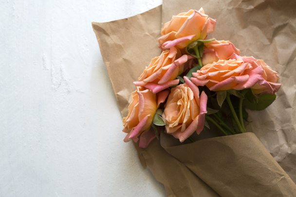 Подарунок, квіти, реміснича упаковка. Помаранчево-рожеві троянди в крафт-папері на білому дерев'яному фоні
 - Фото, зображення