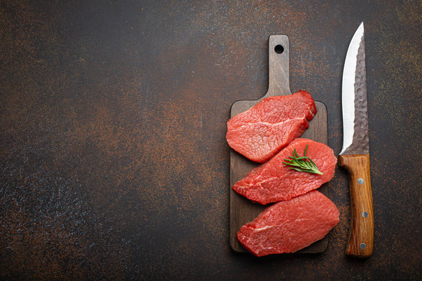 Three raw uncooked meat beef top sirloin steaks on wooden cutting board with rosemary on wooden cutting board with knife on dark rustic background from above, preparing meal with meat, copy space - 写真・画像