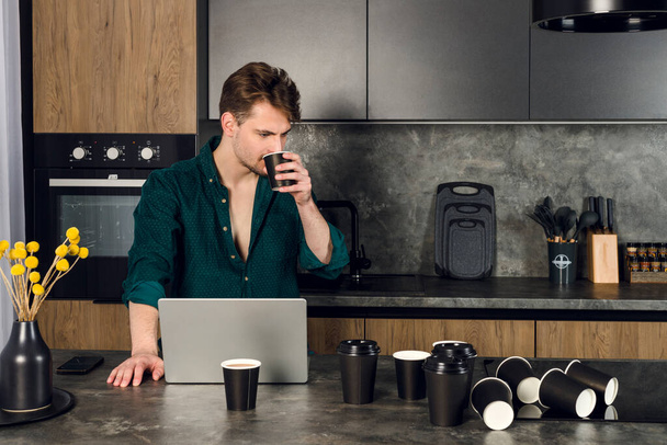 Slaperige man verslaafd aan koffie werken op een laptop in de keuken, veel kopjes verspreid rond. cafeïneverslaving. - Foto, afbeelding