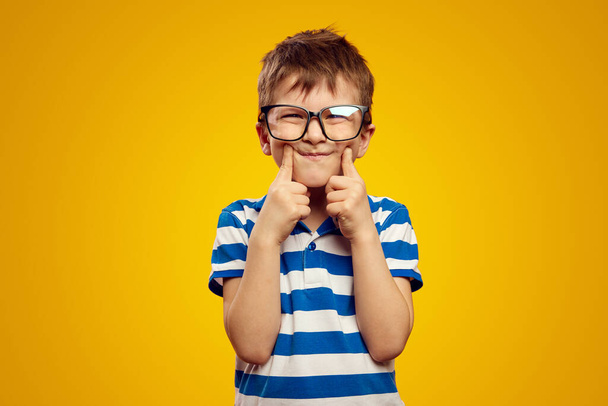 Smart cute kid wearing glasses and blue striped polo, touching cheeks with both index finger against yellow background, making grimace - 写真・画像