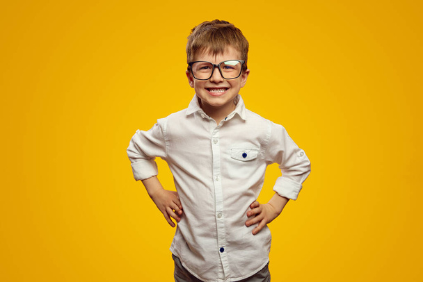 Little nerd kid wearing glasses and white shirt smiling for camera while keeping hands on waist against yellow background - Fotografie, Obrázek