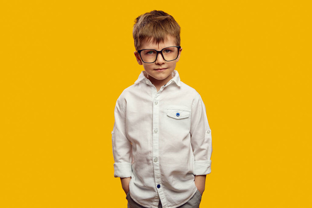 Poważny chłopiec w białej koszuli i okularach, trzymający ręce w kieszeniach i patrzący wściekle na kamerę na żółtym tle - Zdjęcie, obraz