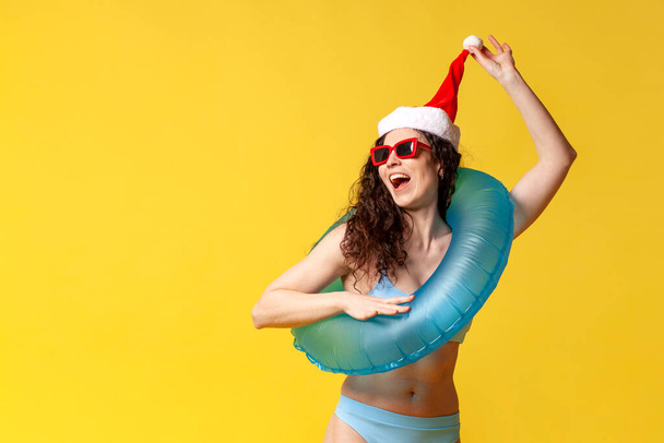geschokt meisje in badpak in santa hoed en met opblaasbare zwemcirkel is verrast op gele achtergrond voor het nieuwe jaar, vrouw gaat op vakantie voor kerst en kijkt naar de kopieerruimte - Foto, afbeelding