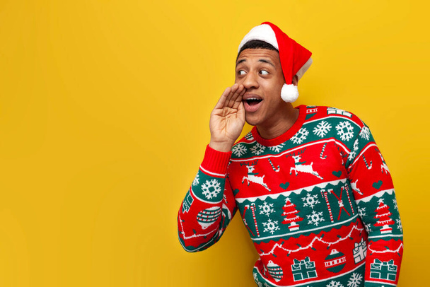 afro-américain gars en vêtements de nouvel an et santa chapeau esquisse et parle sur fond jaune isolé, homme en chandail commérages et raconte secret pour la nouvelle année - Photo, image