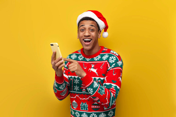 chico afroamericano en ropa de Navidad y sombrero de santa está utilizando el teléfono inteligente y sonriendo sobre fondo aislado amarillo, hombre en suéter está escribiendo en línea en el teléfono para el año nuevo - Foto, Imagen