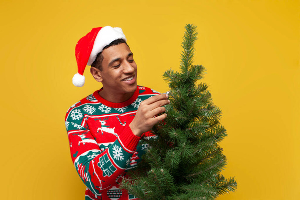 chico afroamericano en ropa de navidad y sombrero de santa sostiene árbol de navidad sobre fondo aislado amarillo, hombre en suéter decora el árbol de navidad para el nuevo año - Foto, Imagen