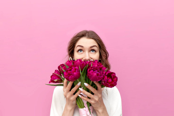 young cute girl in festive clothes sniffs bouquet of flowers and looks up on pink isolated background, woman with pink tulips covers her face with flowers and looks at copy space - Photo, Image