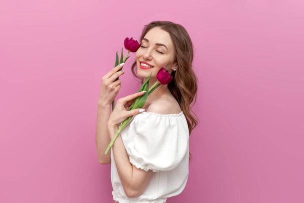 young tender girl touches the skin of her face with two pink tulips and smiles on pink isolated background, woman with closed eyes in white clothes poses with flowers - Photo, Image