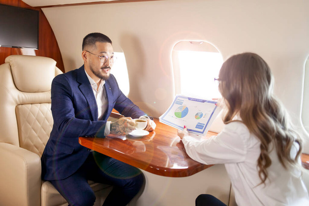 民間航空機で会うビジネスマン訴訟関係のアジア人ビジネスマンは女性の同僚と仕事を話し経営者はグラフを見せ上司に報告するビジネスパートナーはジェットで飛ぶ - 写真・画像