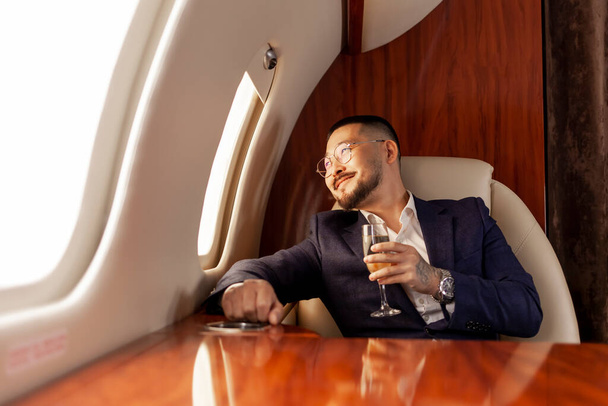 επιτυχημένος ασιάτης επιχειρηματίας με κοστούμι και γυαλιά με ένα ποτήρι σαμπάνια πετάει σε ιδιωτικό τζετ πολυτελείας, Κορεάτης επιχειρηματίας ξεκουράζεται και χαλαρώνει κατά την πτήση, πολυτελή τρόπο ζωής - Φωτογραφία, εικόνα