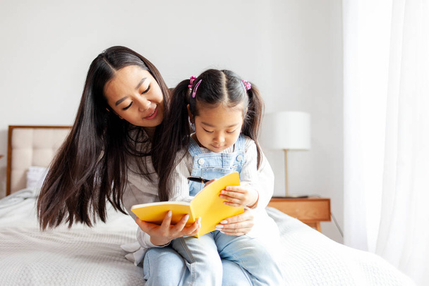 asiatico madre insegna figlia a leggere e scrivere a casa su letto, piccolo coreano ragazza scrive in notebook con genitore, famiglia educazione e educazione - Foto, immagini
