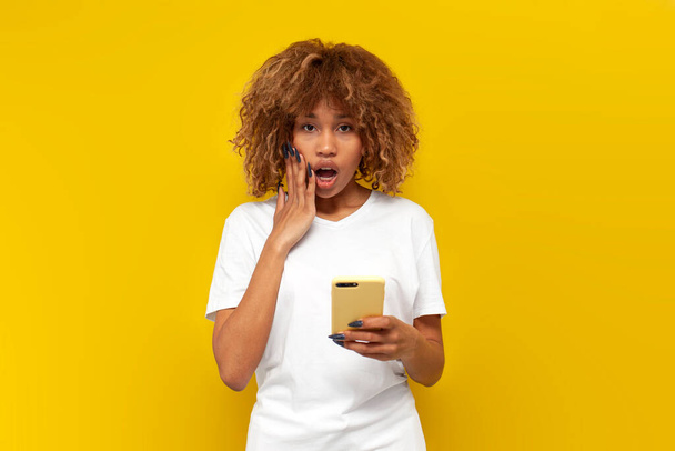 užaslá kudrnatá americká dívka v bílém tričku používá smartphone a divit se na žlutém izolovaném pozadí, šokovaná mladá žena píše zprávu a ukazuje šok - Fotografie, Obrázek